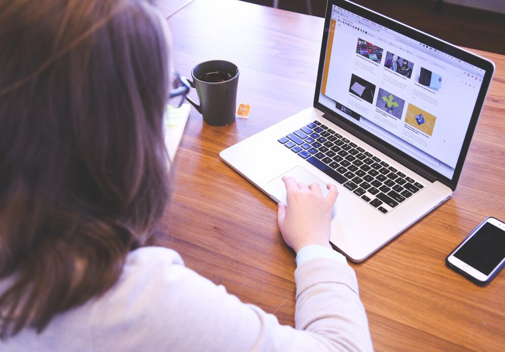 Mujer sentada en una mesa de escritorio, tiene la mano derecha encima del mousepad de la laptop, a la izquierda de la laptop tiene un móvil y a la derecha una taza de café.