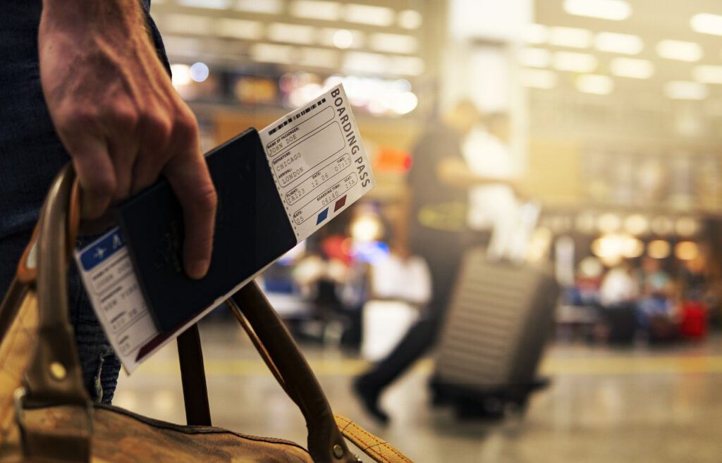 Primer plano una mano de una persona sosteniendo una valija y a la vez un pasaporte con el boleto de viaje, de fondo luces y personas en un aeropuerto.