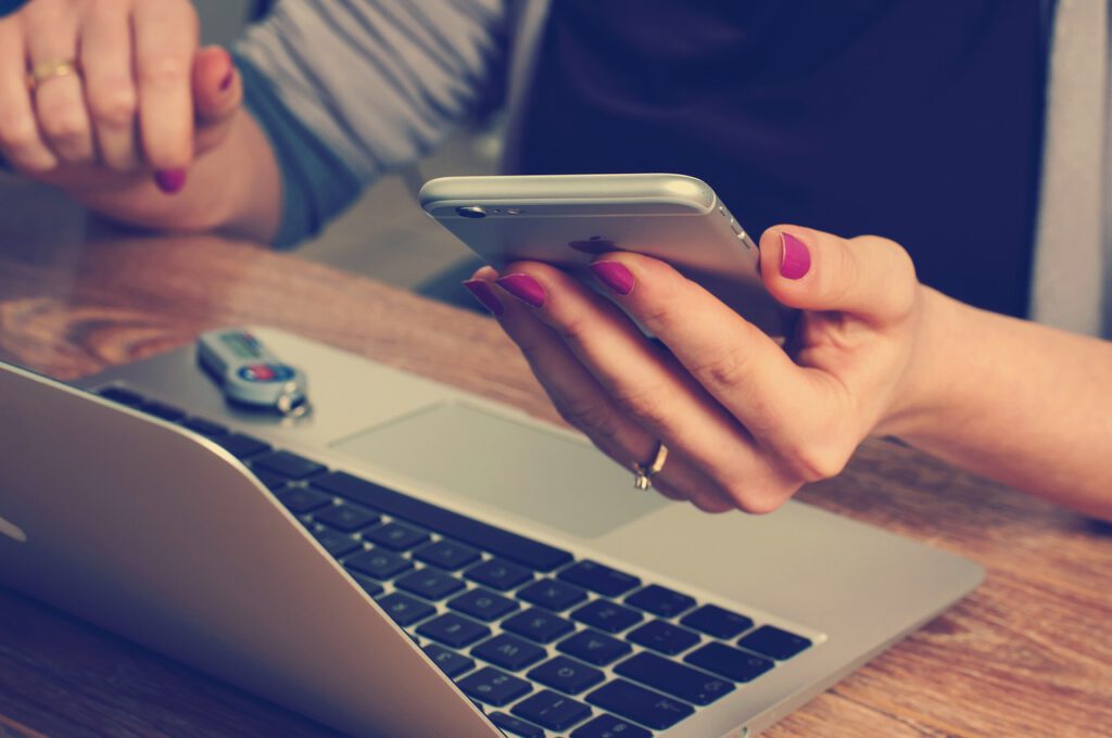 Manos de una mujer con un teléfono móvil en la mano, sentada en un escritorio con una laptop abierta 